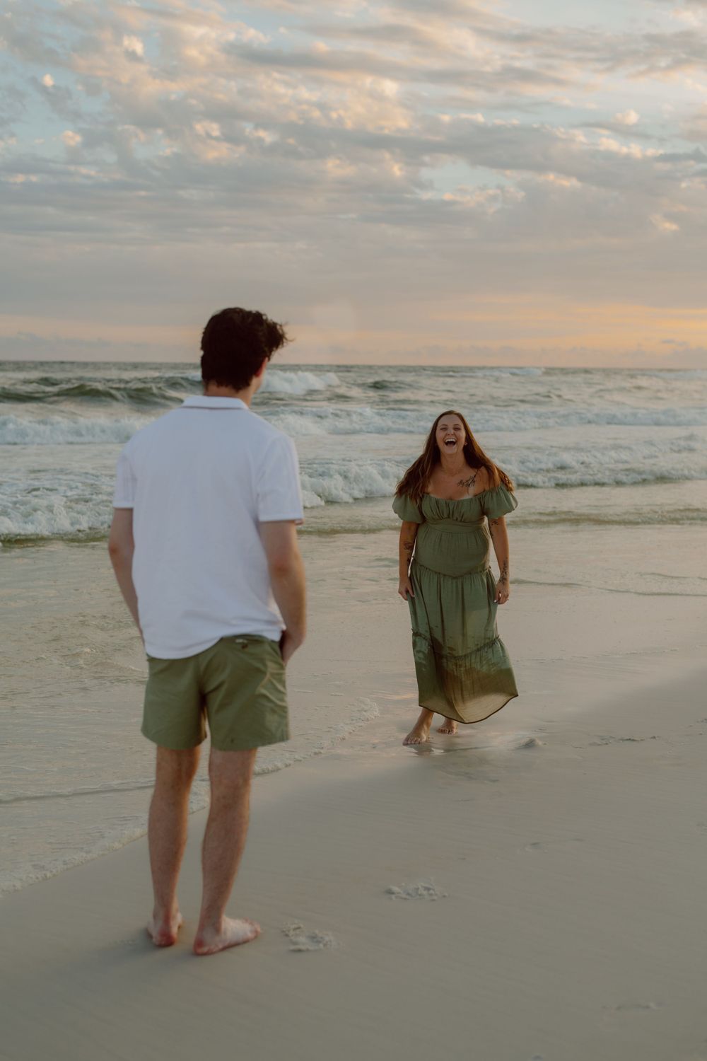 A couple stands on a beach at sunset with crashing waves and cloudy skies in the background.