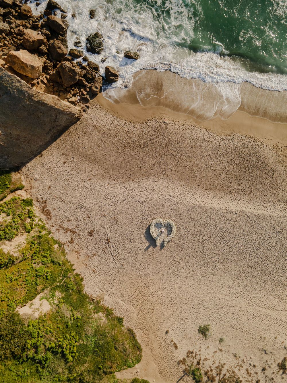 Aerial view of a heart shape drawn in the sand on a pristine beach next to rocky cliffs.