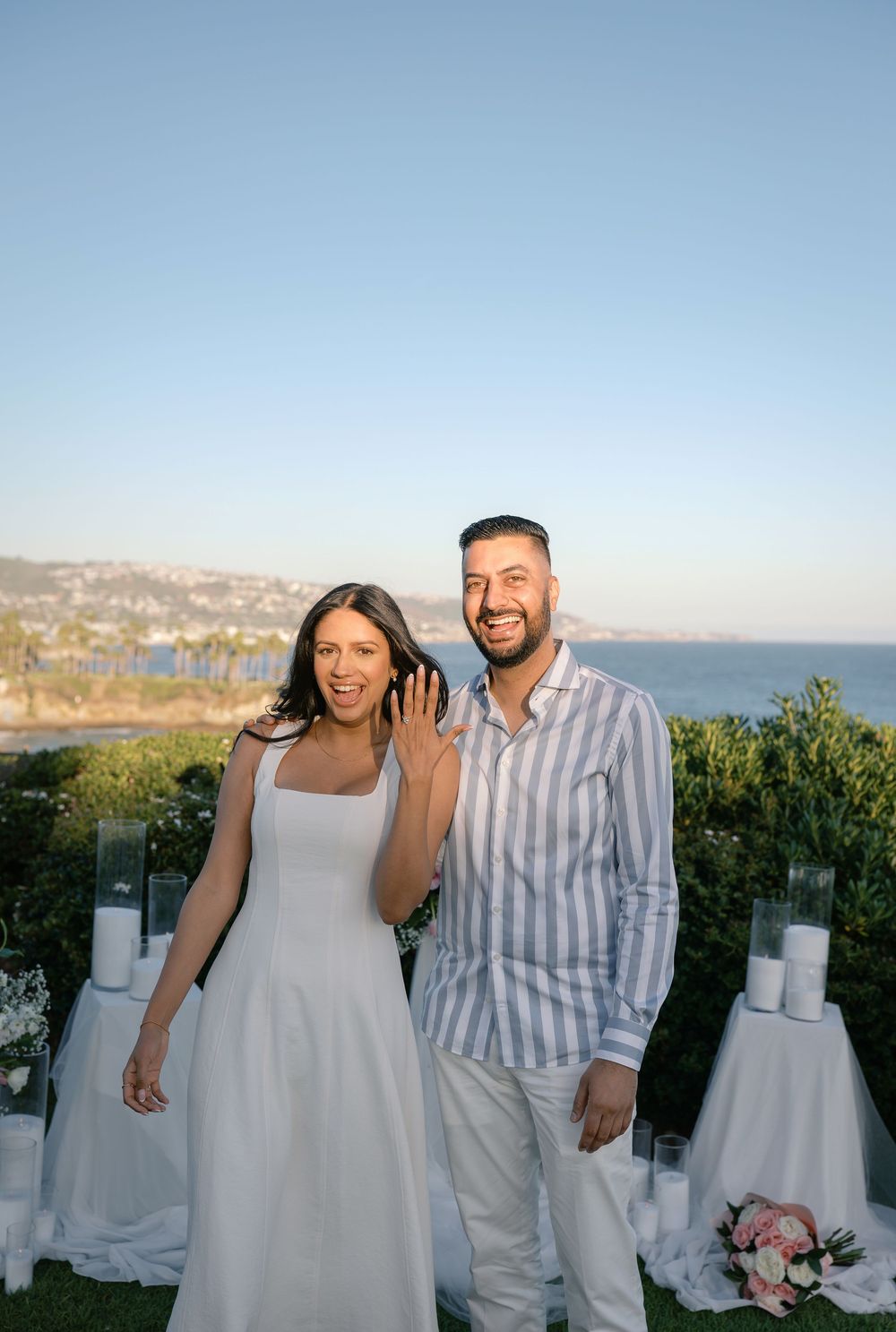 Happy couple poses outdoors at sunset near the ocean, woman showing engagement ring, with candles and flowers in background.
