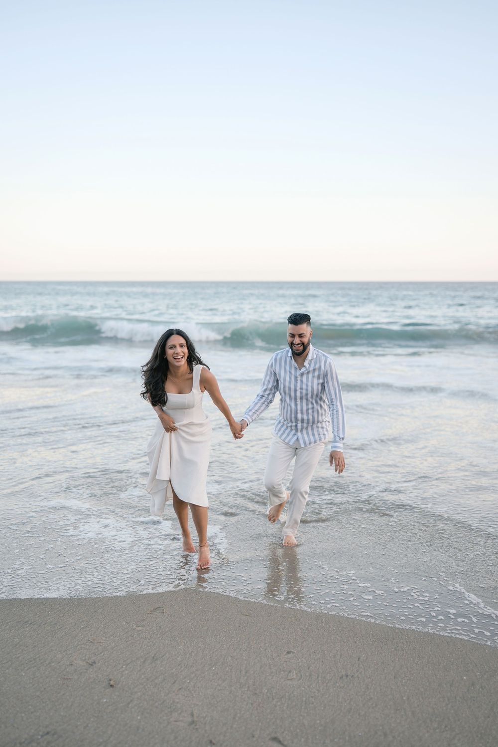 A joyful couple holds hands and walks along the shoreline at sunset, wearing white outfits with waves behind them.