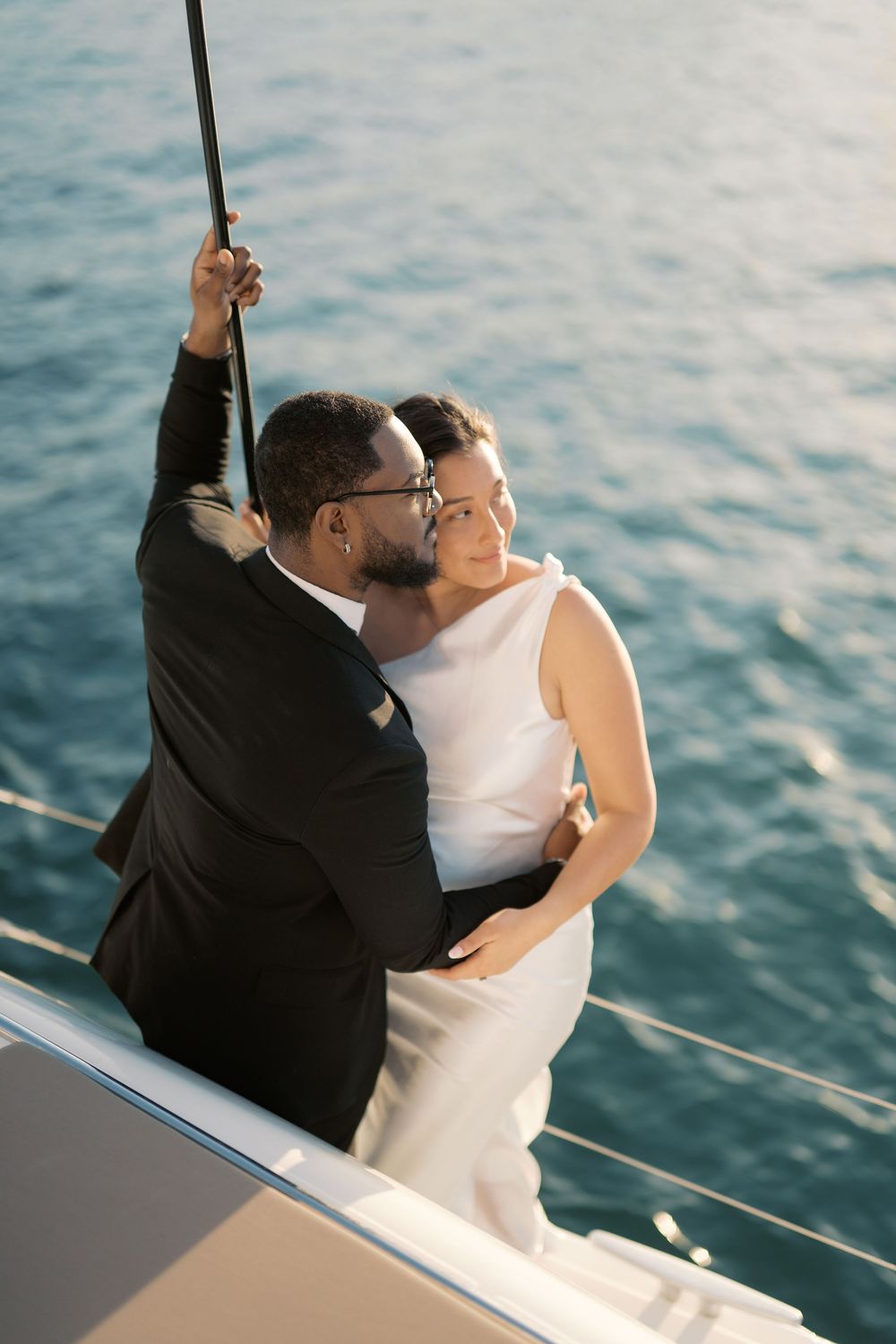 Interracial couple embracing romantically on a yacht deck, groom in black suit and bride in white dress with ocean backdrop.