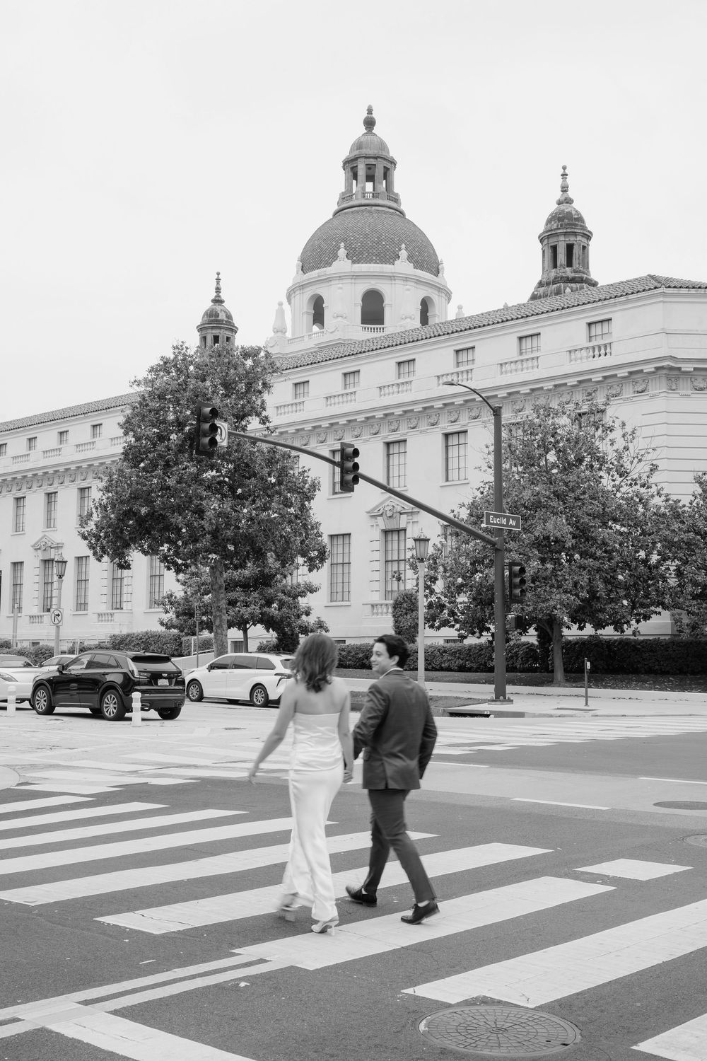 Black and white photo of a couple in wedding attire crossing a street in front of a grand domed building.