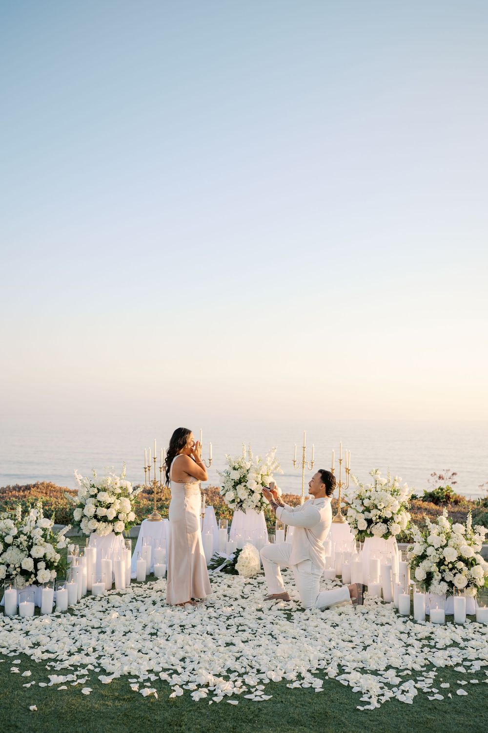 Man proposes on one knee amid white flowers and candles with ocean backdrop at sunset.
