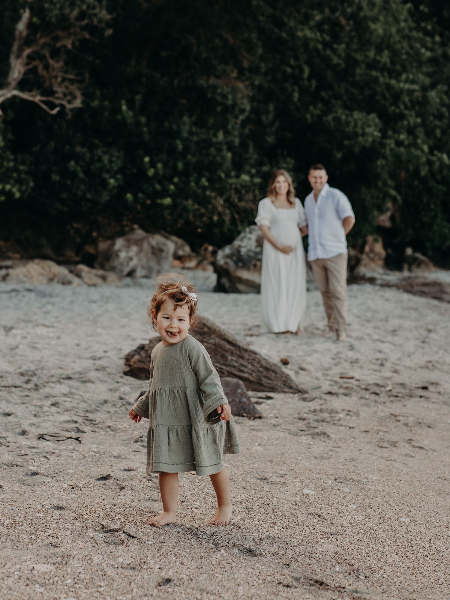 Family beach photo with toddler in green dress playing in foreground on sandy shore.