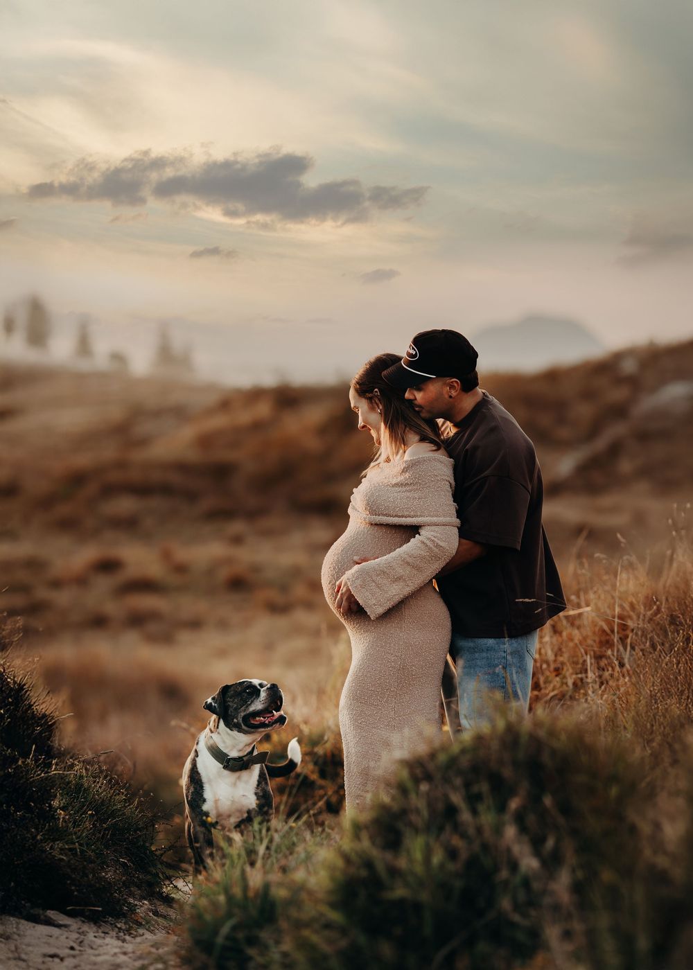 A couple shares a tender moment on a hillside at sunset with their dog sitting beside them in the golden light.