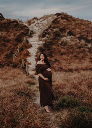 A figure in a black dress walks along a winding path through rustic brown coastal terrain at sunset.
