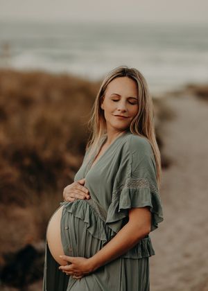 An expectant mother in a sage green flowy dress stands on a beach at golden hour, tenderly cradling her baby bump.