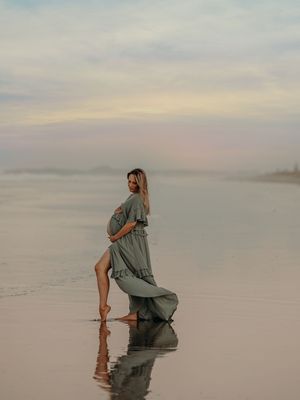 A pregnant figure in a flowing sage green dress poses elegantly on a misty beach at sunset, creating reflections in the wet sand.