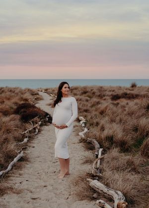 A figure in a white dress walks along a sandy coastal path through dried grass at sunset.
