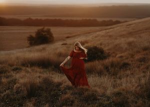 A figure in a flowing red dress stands on a golden sunlit hillside at dusk while their hair blows in the wind.