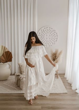 Woman wearing a white off-shoulder tiered maxi dress poses in a boho-style room with neutral decor and pampas grass.