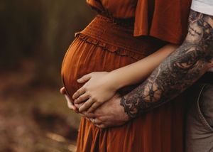 Close up of expecting couple with person wearing burgundy dress cradling baby bump while standing outdoors.