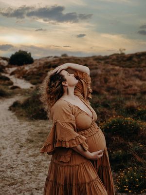 A person in a flowing brown dress stands on a sandy path at sunset against a dramatic cloudy sky.