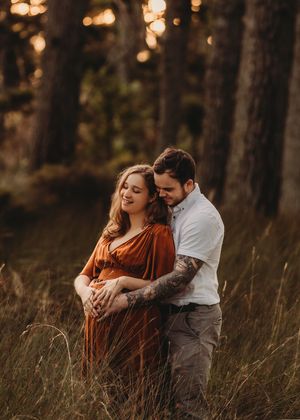 A couple embraces in a sunlit forest clearing, with golden hour light filtering through tall trees around them.