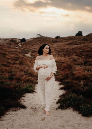 Ethereal series of desert maternity photos showing a woman in white dress walking on sandy dunes at sunset.