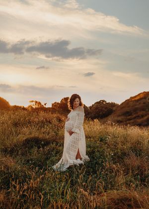 A figure in a flowing white lace dress poses in a golden field at sunset with rolling hills in the background.