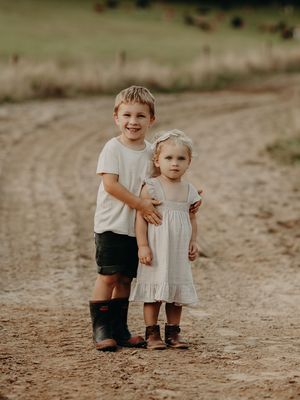 Two children stand together on a dirt path wearing boots, with one in a white dress and the other in shorts.