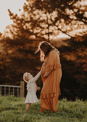 Mother and child holding hands in a golden sunset glow while standing on grassy hill in matching light dresses.