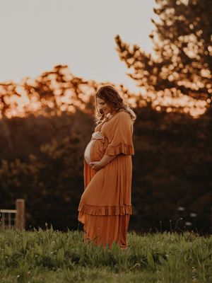 An expectant mother in a flowing rust-colored dress stands in a field at sunset, cradling her baby bump.