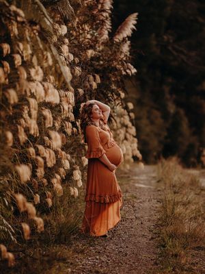 Elegant figure in a flowing rust-colored dress stands on a path against a rustic stone wall with autumn foliage above.
