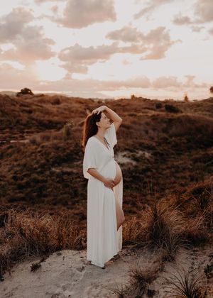 A couple in white flowing dresses embrace on a sandy hill during sunset creating a romantic silhouette.