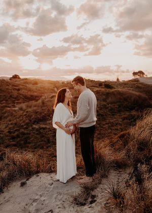 A couple stands together on a hilltop during golden hour sunset with warm tones and dramatic clouds in the sky.