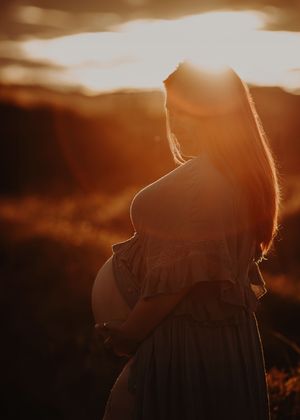 A silhouette of a pregnant woman standing in warm golden sunset light with a dramatic atmospheric sky.