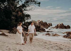 A couple in white outfits walks along a scenic beach with rocky cliffs and ocean waves in the background.