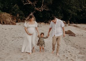 Family holding hands while walking on beach, showcasing expectant mother in white dress.