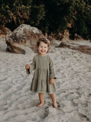 A small child stands barefoot on a sandy beach wearing a tiered olive green dress while laughing joyfully.
