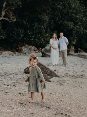 A young child in an olive dress plays on a sandy beach while two adults stand in the background.