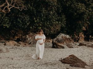 A woman in a flowing white dress stands on a sandy beach surrounded by large rocks and greenery.