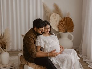 A couple in warm earth tones embrace intimately in a minimalist room with dried palm fronds and neutral decor.