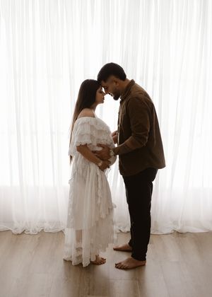 An expectant couple shares an intimate moment against white curtains in a maternity photo shoot.