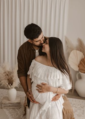 Romantic maternity photo of an expectant couple embracing in a white room with neutral decor and dried flowers.