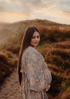 A woman in a patterned blouse poses gracefully against a golden sunset on a hillside trail during golden hour.