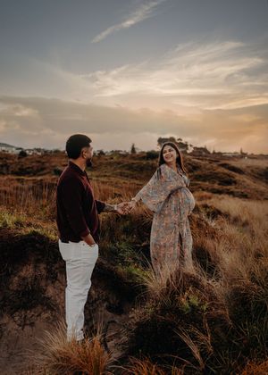 A couple holds hands on a hilltop during golden hour, with the woman wearing a flowing floral dress against sunset sky.