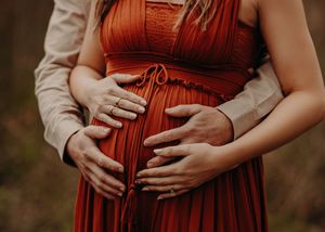 Hands lovingly cradle a pregnant belly in a flowing rust-colored maternity dress against a dark outdoor backdrop.