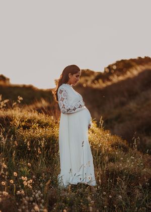 A pregnant woman in a flowing white lace dress stands in a golden field at sunset.