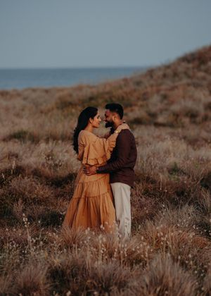 A couple in warm earth tones embraces in a desert landscape with dry brush and golden light at sunset.
