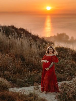 A pregnant woman in a flowing red dress stands on a cliff against a stunning orange sunset over the ocean.