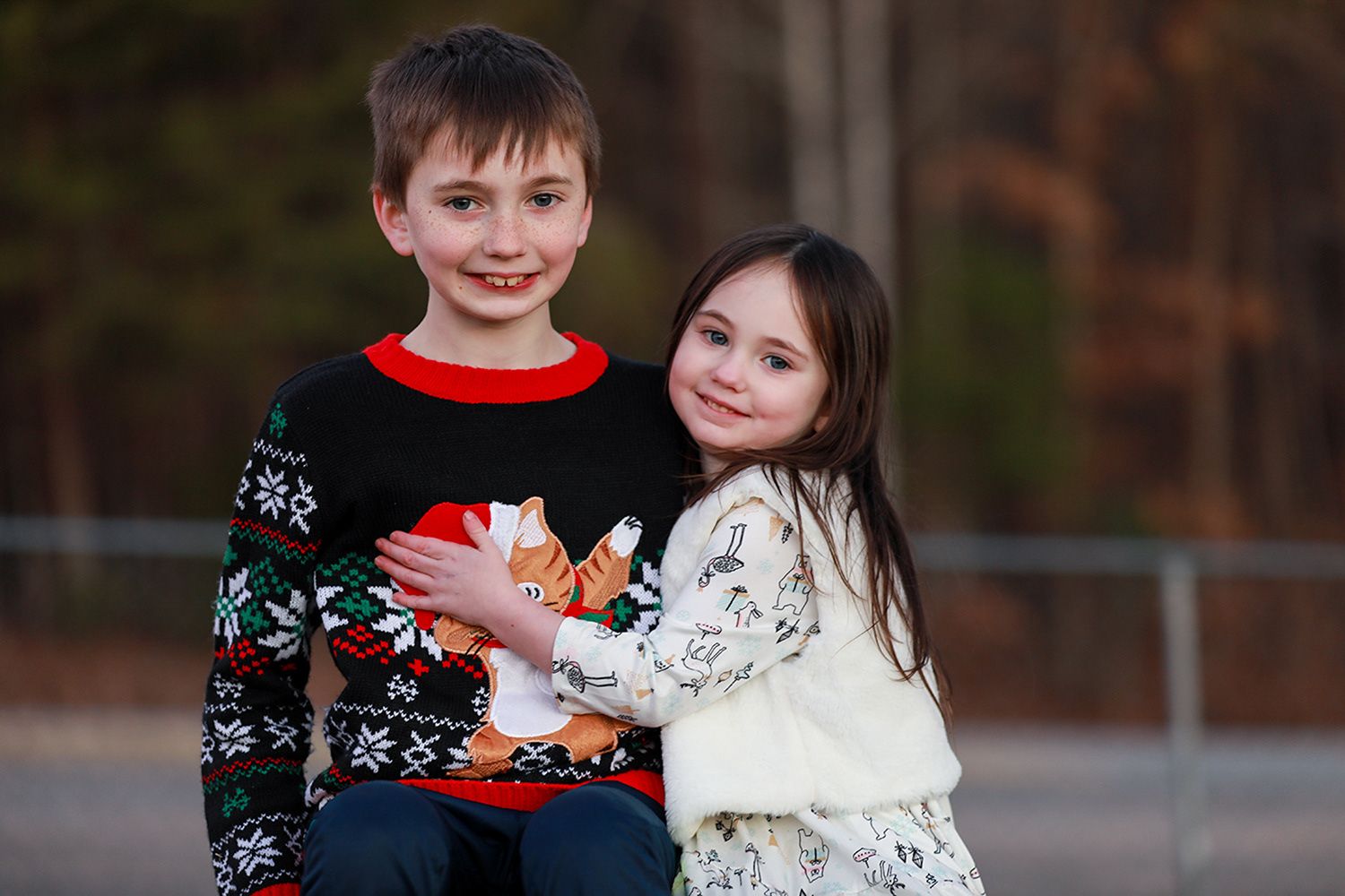 Two children in holiday sweaters pose together outdoors in a warm autumn setting.