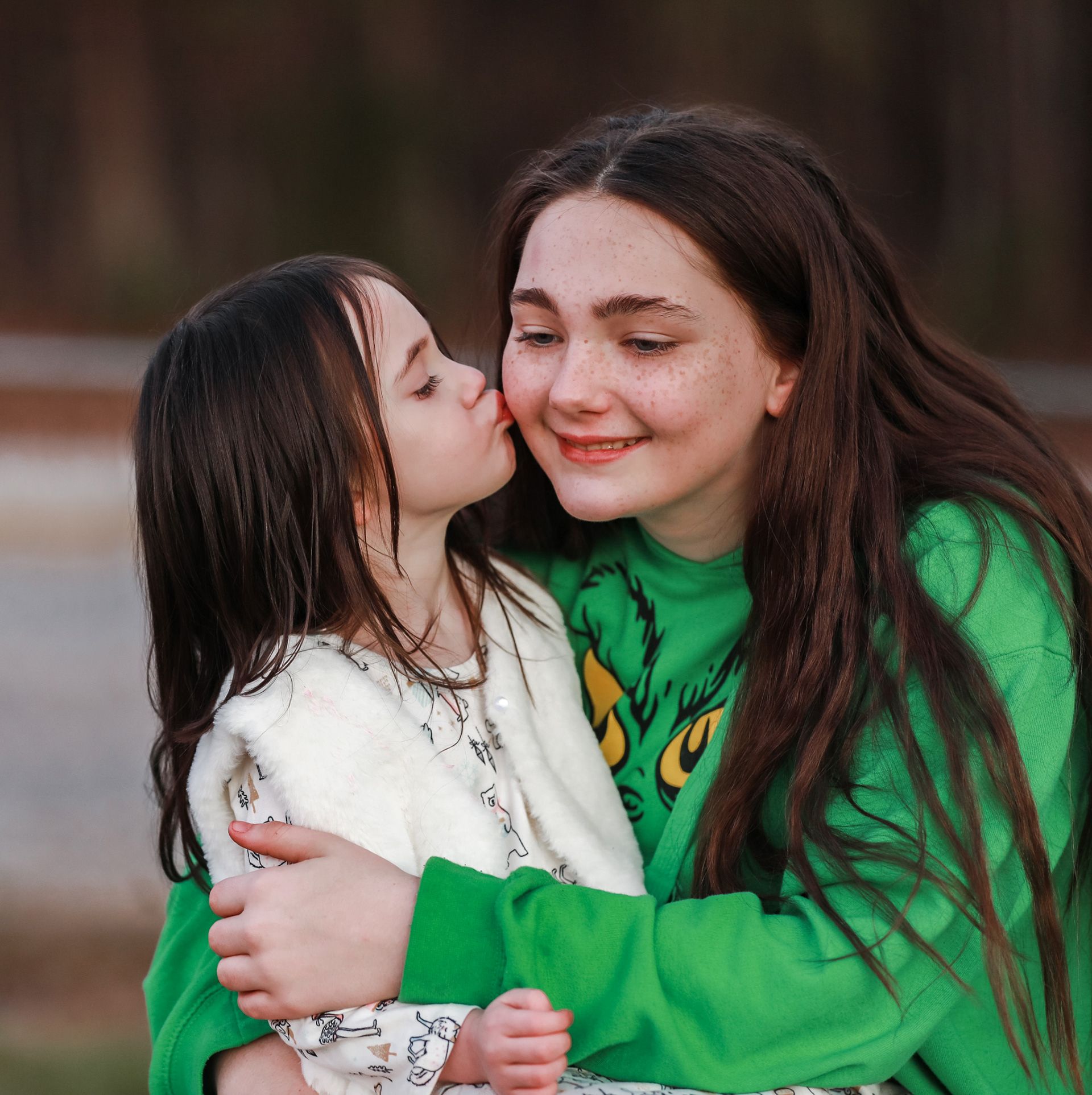A young girl kisses a smiling teenage girl in a green sweatshirt outdoors, sharing a sweet tender moment.