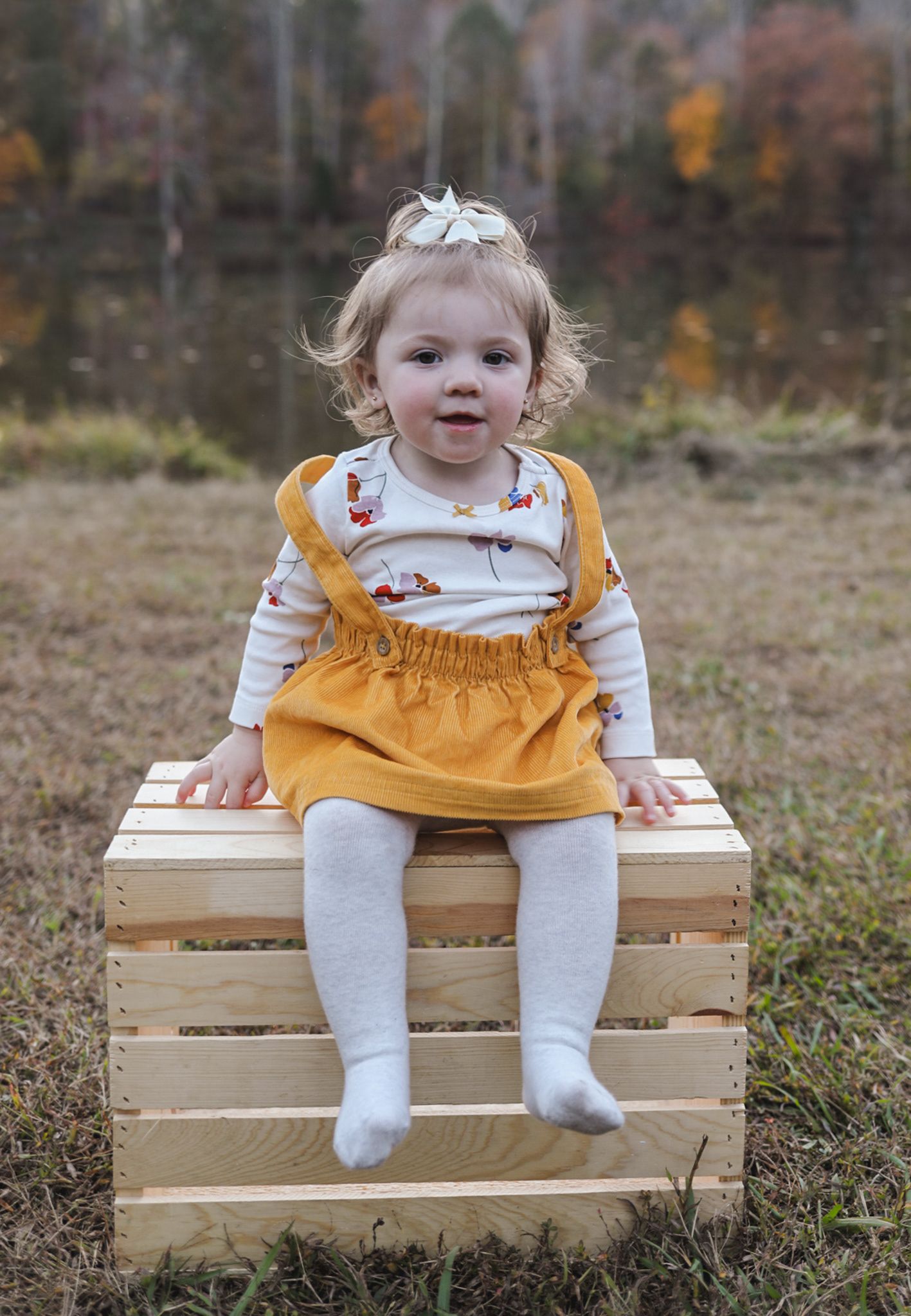 Adorable toddler girl in mustard skirt sitting on a wooden crate in an autumn outdoor setting.