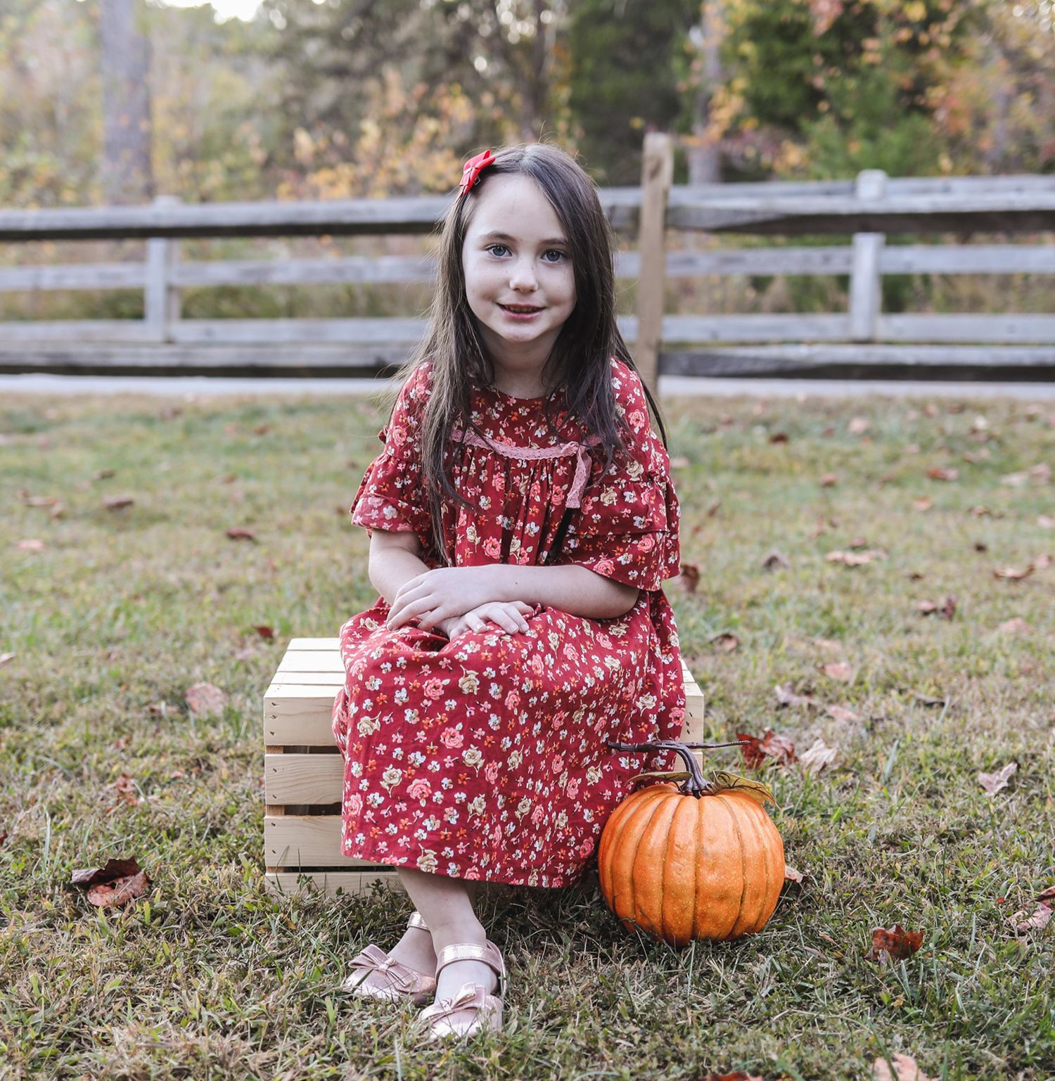 Young girl in red floral dress sits on wooden crate beside orange pumpkin outdoors in autumn setting.