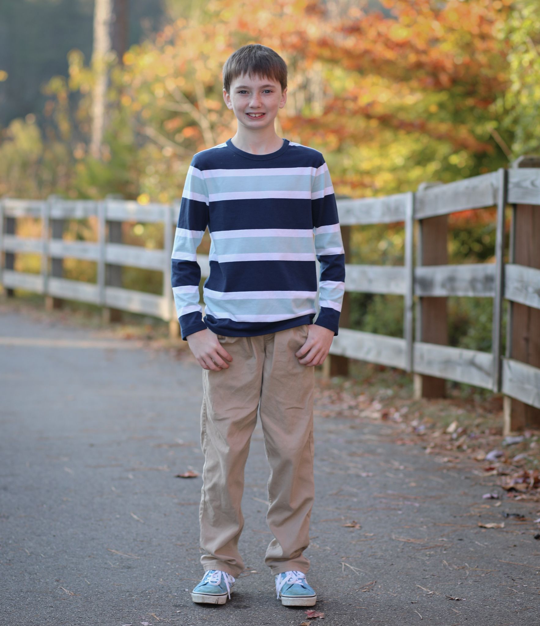 Smiling boy in striped navy shirt and khaki pants standing on a path with autumn foliage background.