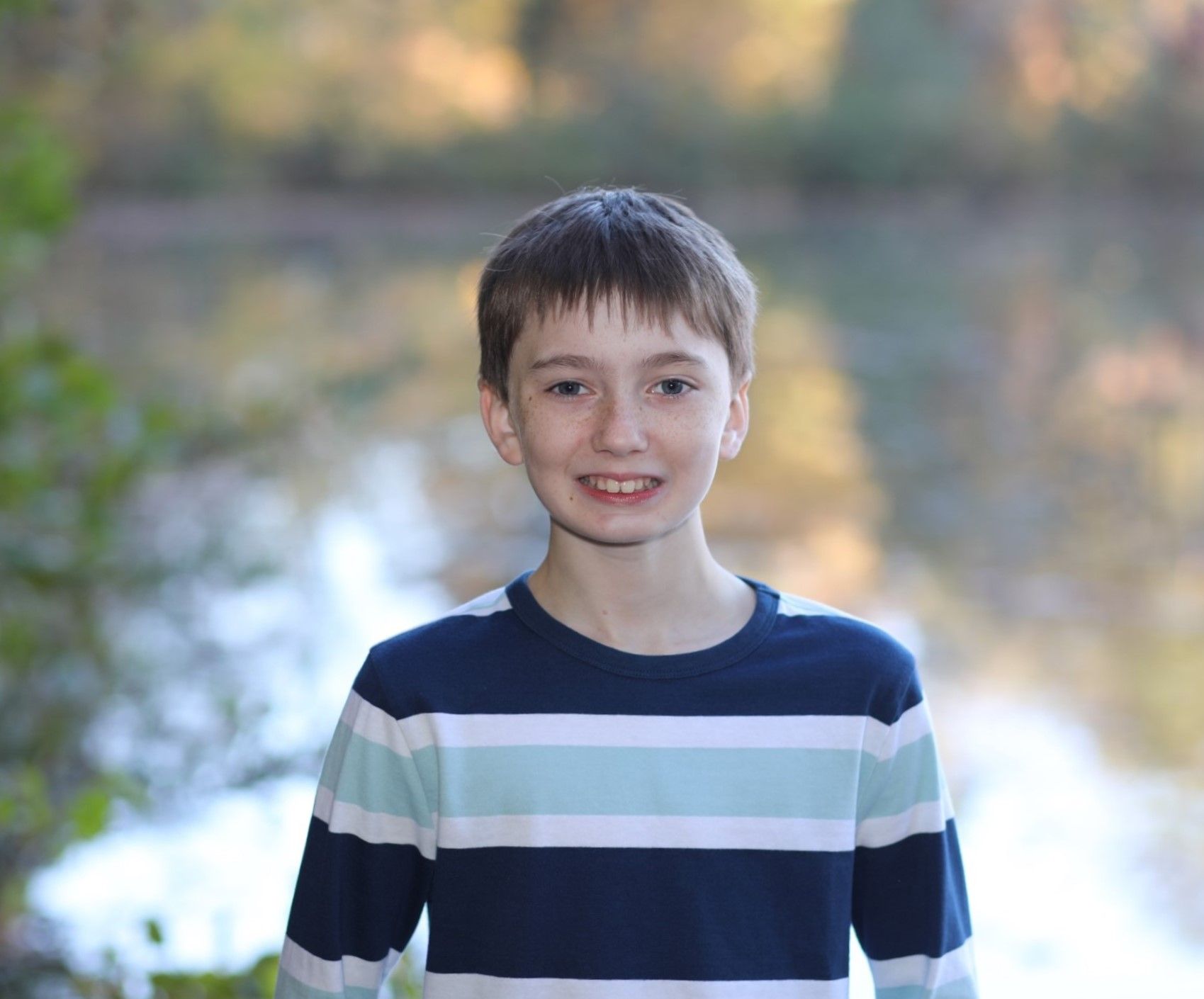 Smiling young boy in striped navy shirt posing outdoors near a pond with blurred autumn foliage background.