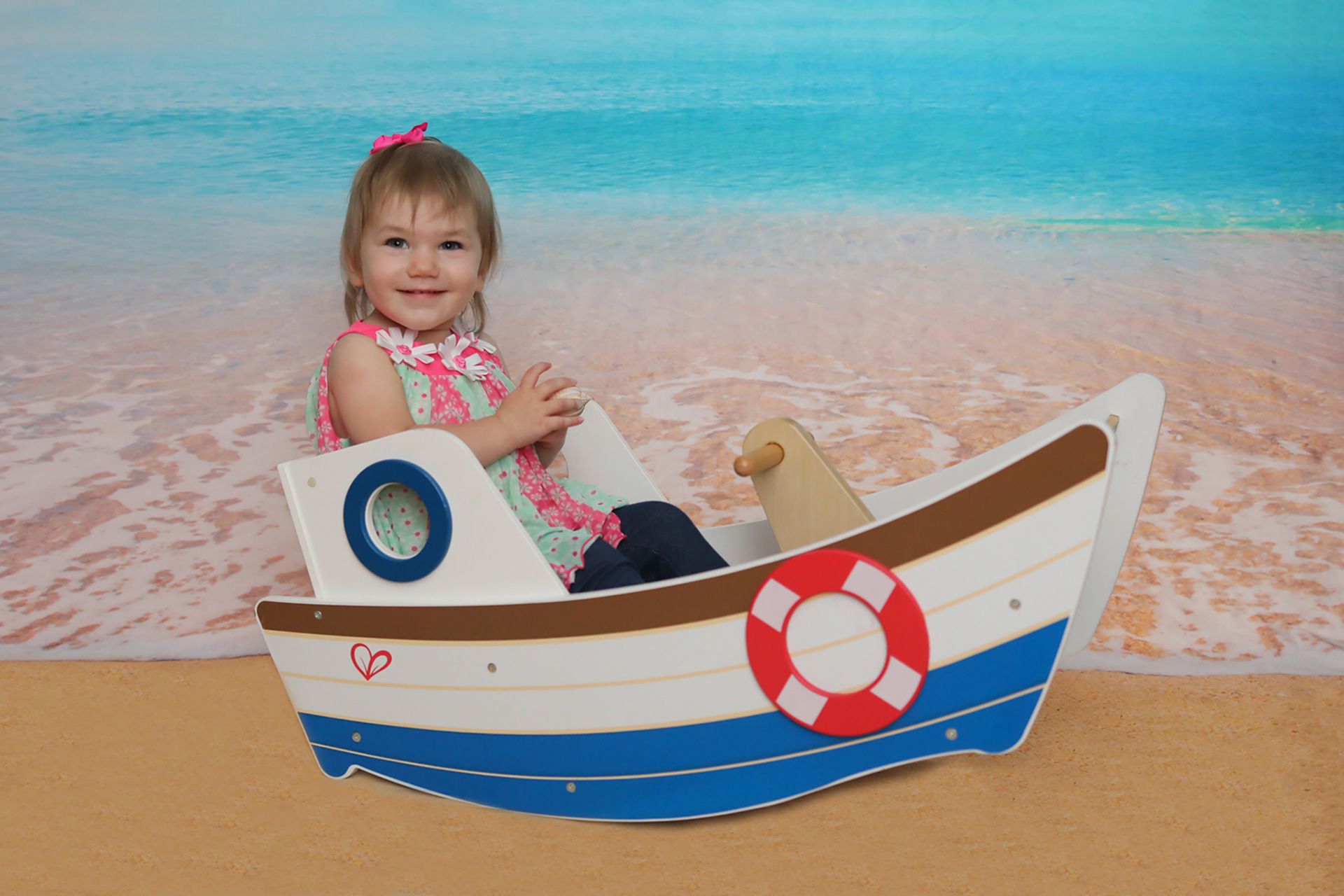 Toddler girl smiling while sitting in a colorful wooden toy boat with a life preserver, set against a beach backdrop.