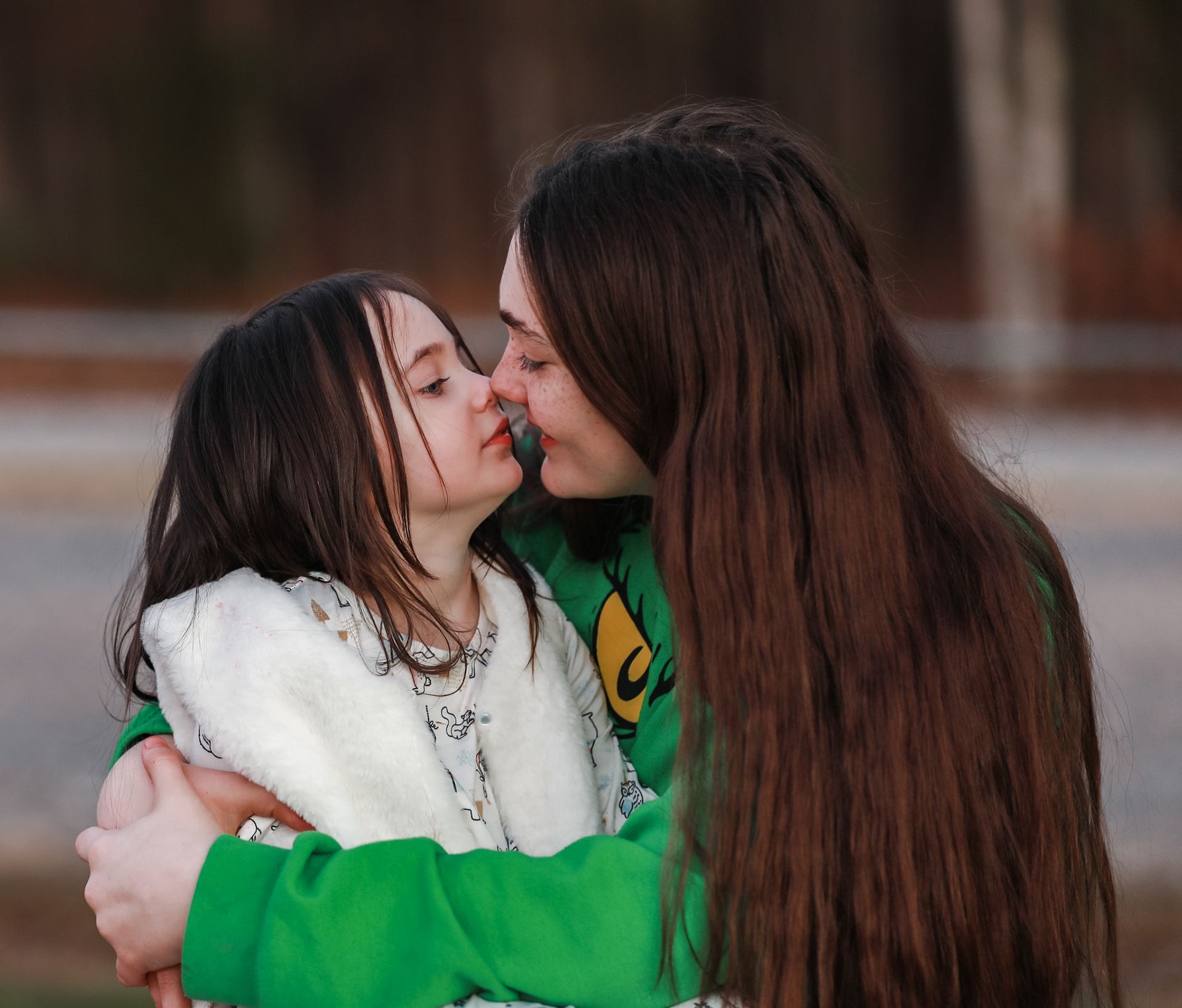 Two young women sharing a tender kiss outdoors, one wearing a green jacket and the other in a white fluffy coat.