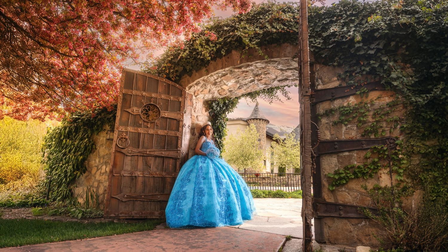 Young woman in blue ball gown quinceañera dress standing under stone archway with autumn foliage in garden setting.